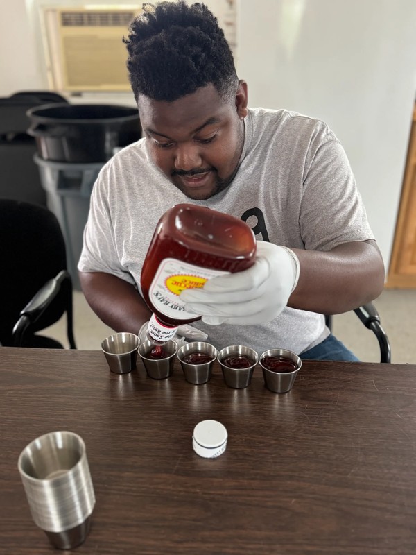 Adult with disabilities pouring ketchup into small metal serving cups during food preparation training at Ivey Ranch ADP