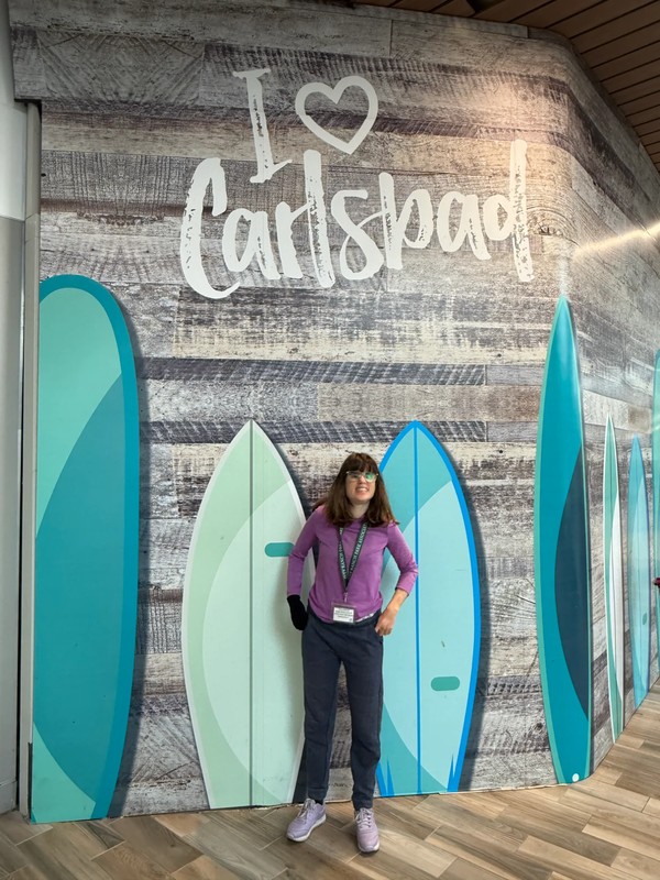 Adult day program participant standing in front of I Love Carlsbad sign with surfboard decorations during community outing