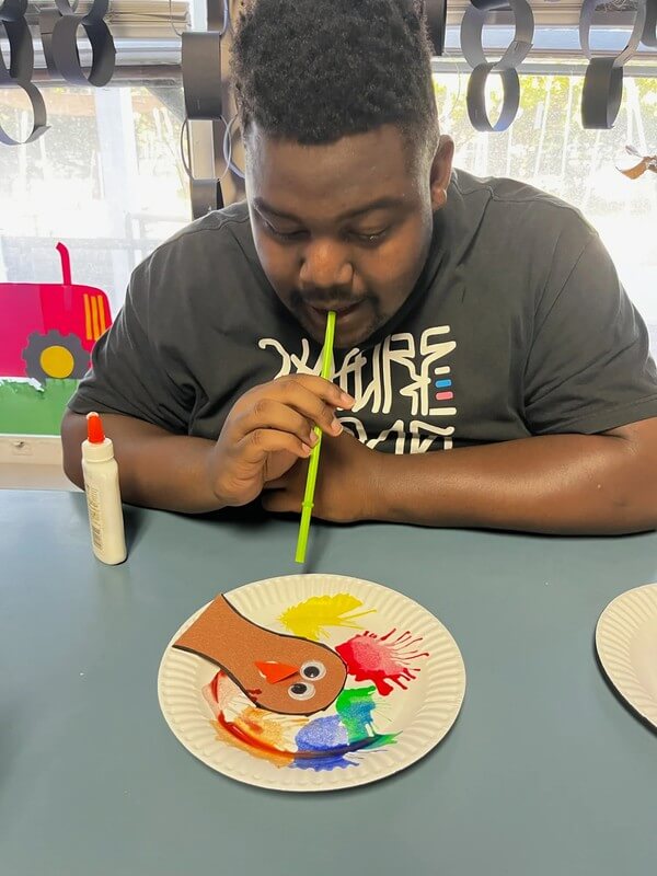 Participant doing art project on a paper plate with a turkey head. He is blowing paint onto the plate for the turkey's feathers.