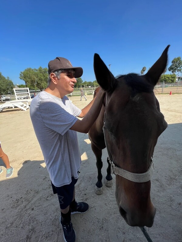 Participant wearing sunglasses gently petting brown therapy horse outdoors at ranch