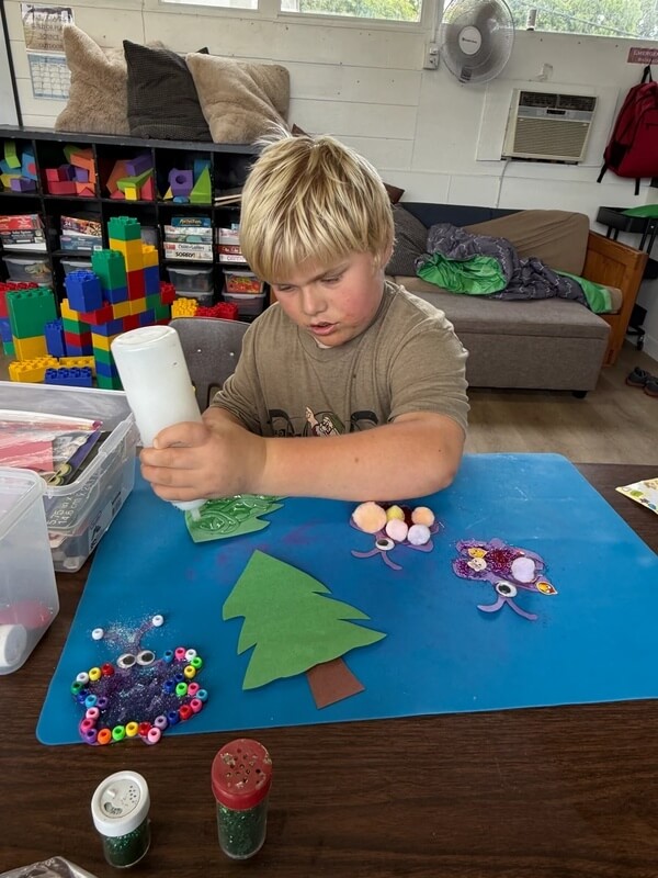 School-age child applying glue to craft project with colorful beads and glitter at table during free play arts and crafts time