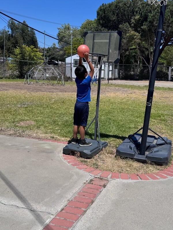 Young boy in blue shirt shooting basketball at portable hoop on outdoor playground court during school age program