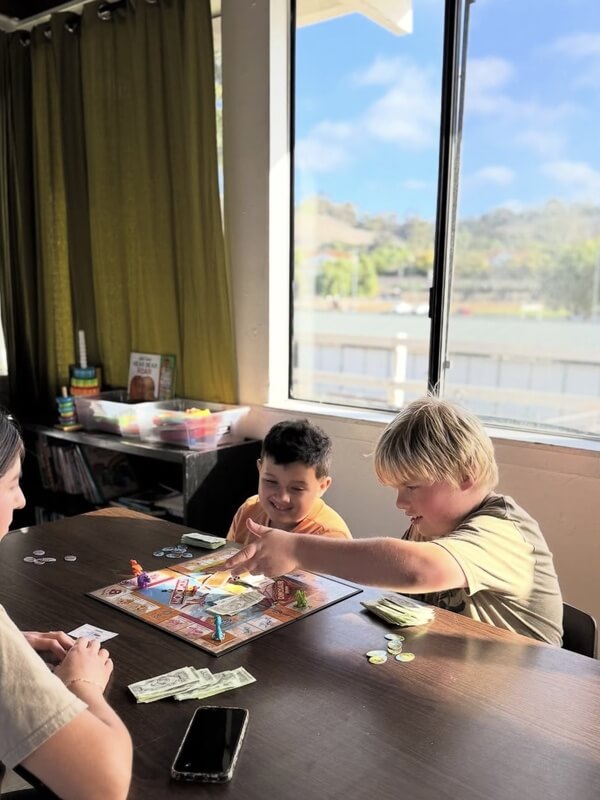 Two children enjoying board game with play money at sunny window table during school age program indoor play