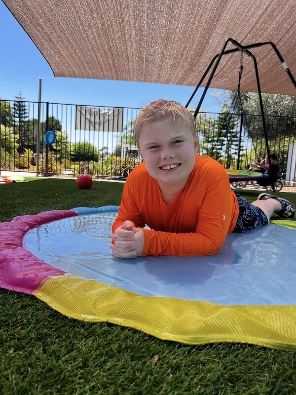 Child relaxing on colorful water play mat during summer camp activities, engaging in sensory experiences and outdoor water fun at daycare program.