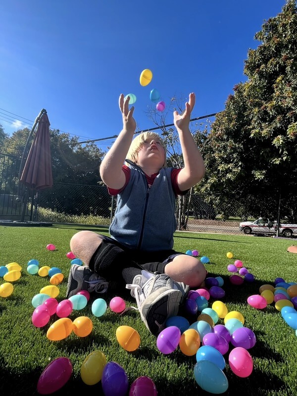 Child surrounded by colorful plastic eggs during outdoor play activity at school-age program, engaging in seasonal games and coordination exercises.
