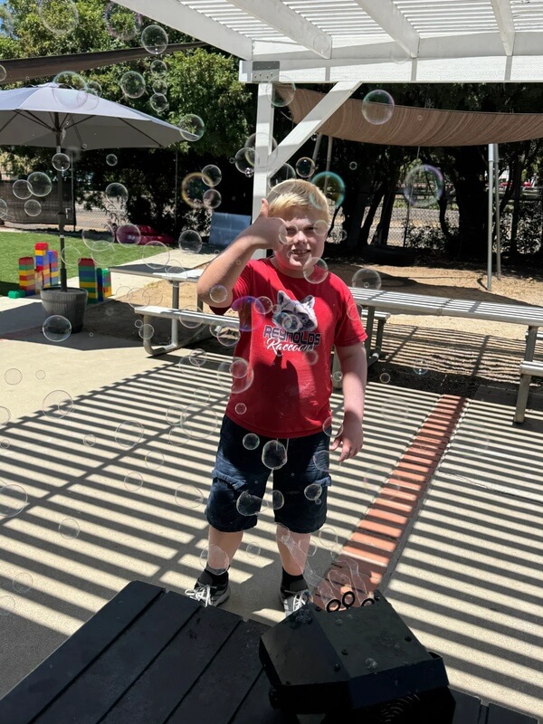 Happy child in red shirt blowing bubbles on shaded outdoor deck with colorful blocks visible in background