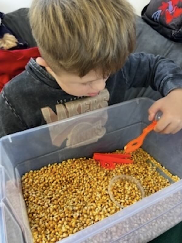 Boy engaged in tactile sensory play with dried corn bin at school-age program, promoting hands-on learning and sensory development through exploration.