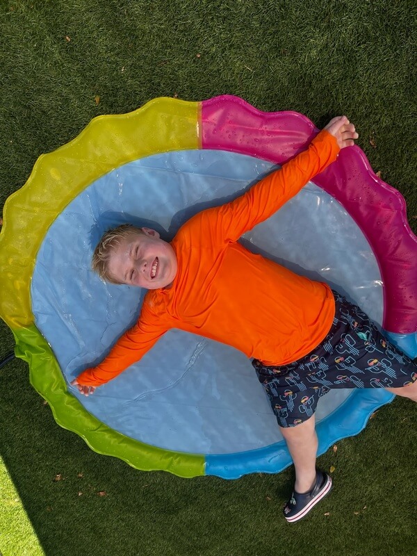 Smiling child in bright orange shirt relaxing in rainbow colored inflatable pool filled with water on grass
