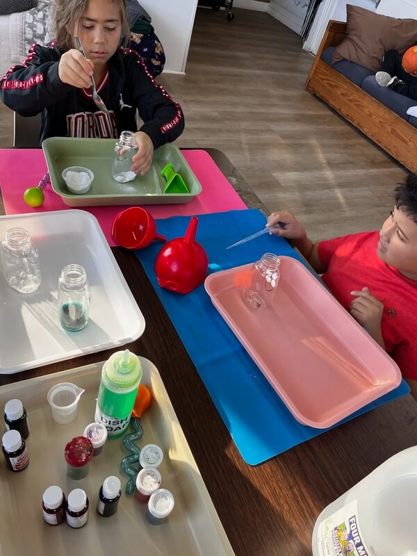 Two children sitting at a table creating colorful sensory jars with glitter, food coloring, and small bottles during hands-on science activity
