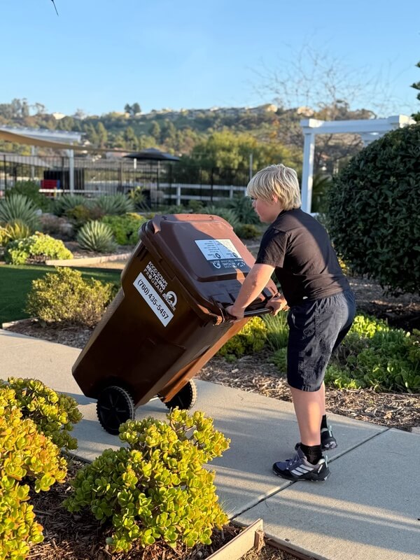 Boy pushing brown trashbin on pathway demonstrating environmental responsibility and waste management skills at after school program