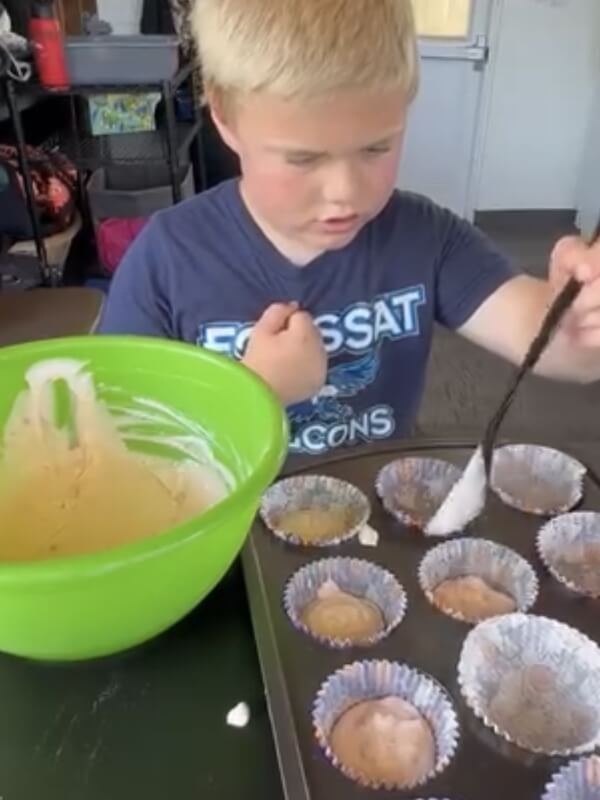 Young boy carefully spooning batter from green mixing bowl into muffin tin liners during baking project