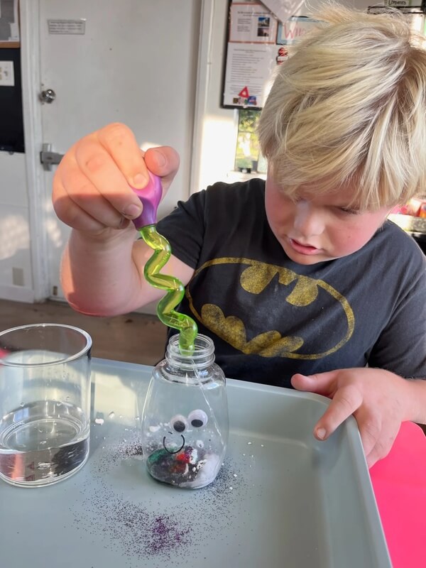 Young boy using funnel to add green glitter into small jar decorated with googly eyes during sensory play activity