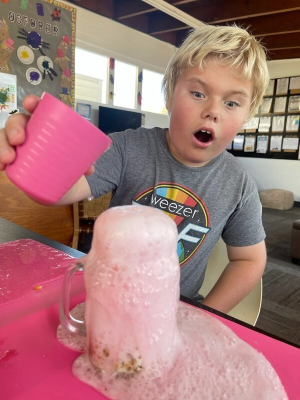 Excited child pouring liquid into jar creating dramatic pink foaming overflow reaction during hands-on chemistry experiment