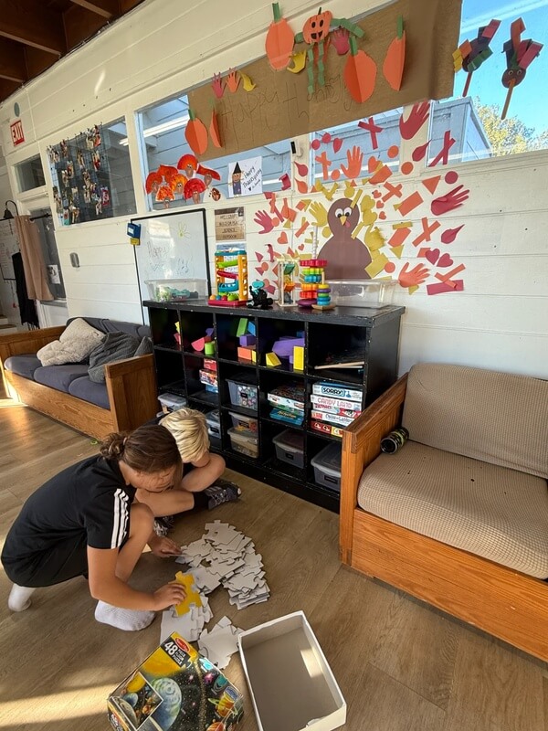 Children on floor assembling large cardboard puzzle pieces near storage shelves with fall decorations on classroom wall