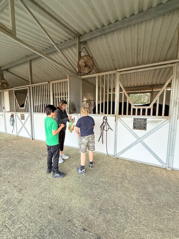 Two children with staff feeding treats to horses through stable stall windows named Lenny during animal interaction time