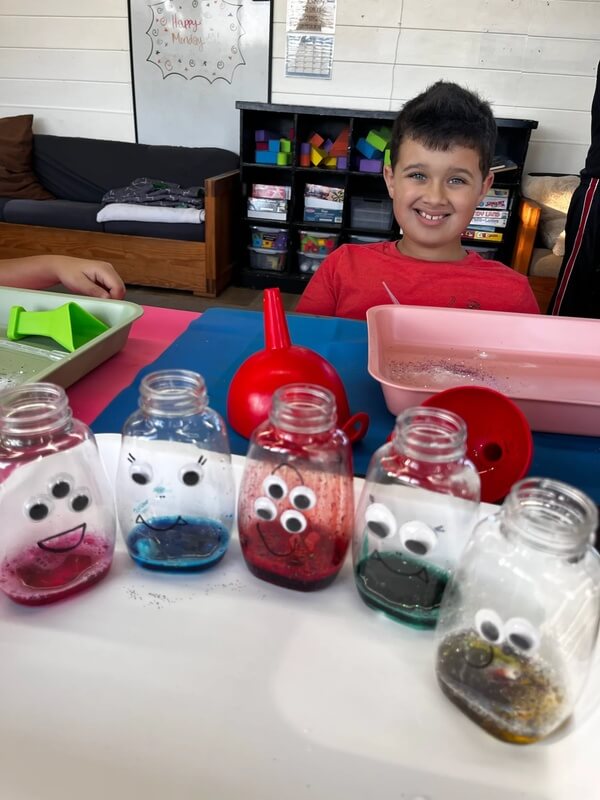 Child displaying five colorful sensory bottles with googly eyes and drawn faces, containing glitter water in blue, pink, red, and green