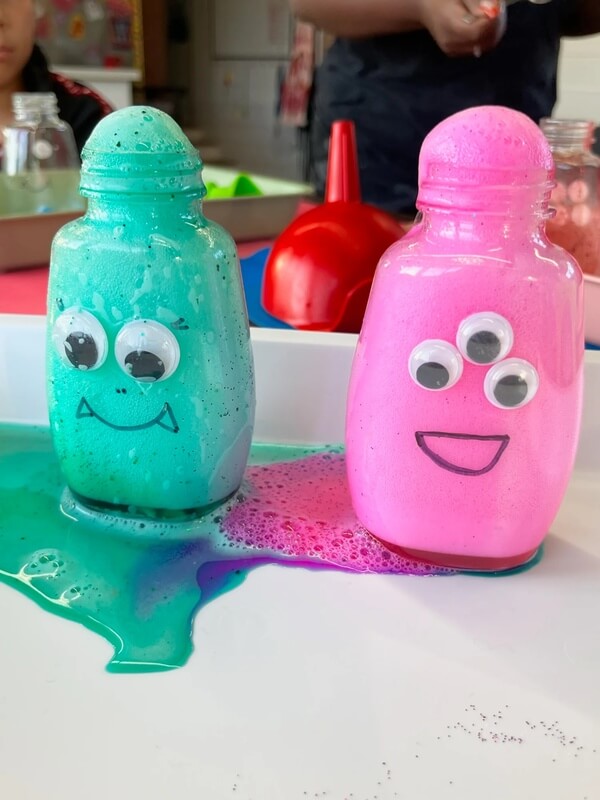 Two glittery sensory bottles in turquoise and pink with googly eyes and smiling faces, showing sparkly liquid contents on table