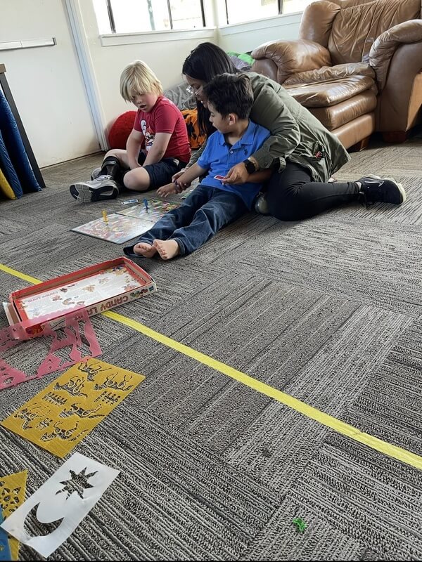 Three children seated on carpet playing collaborative floor game with game boards and yellow tape markers in comfortable classroom setting
