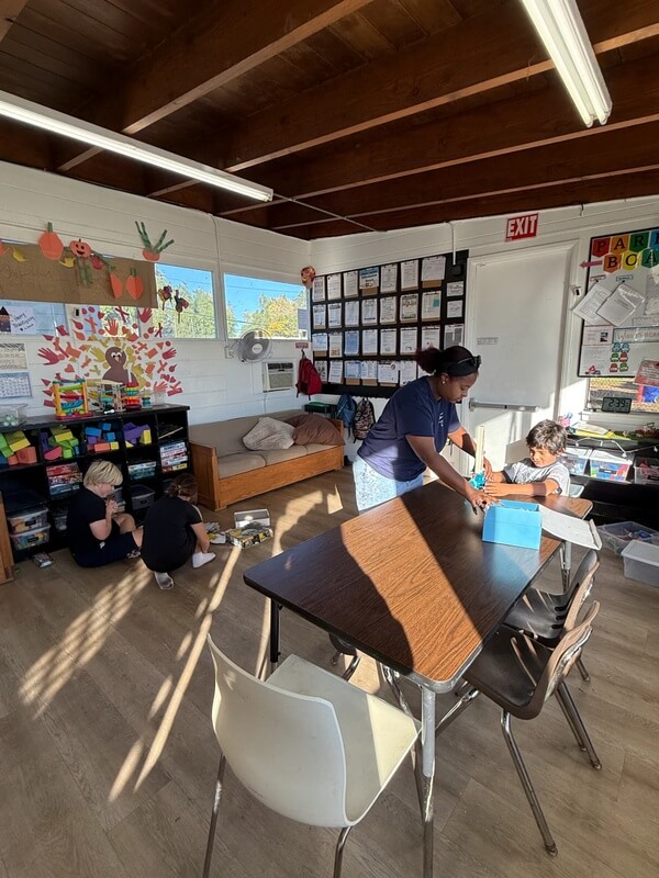 Staff member helping children with activities at table while others play independently in bright classroom with fall decorations and organized storage