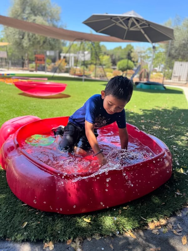 Young child in dark shirt playing and splashing in red inflatable pool on sunny day at outdoor play area
