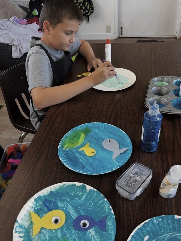 Child gluing fish cutouts onto painted ocean paper plates during sea life themed craft activity