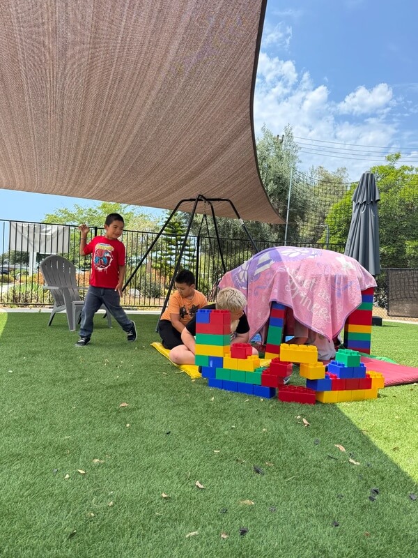Three children constructing colorful fort structure with large foam building blocks under shade sail on artificial turf playground