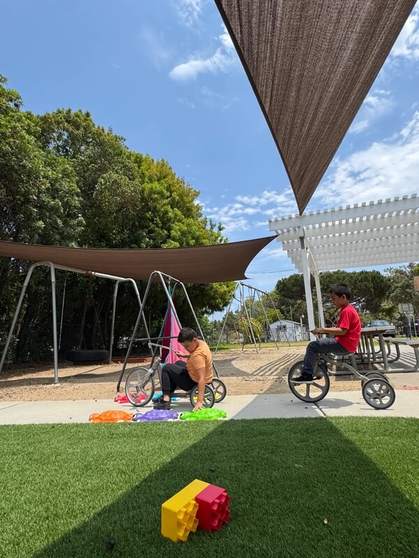Children riding tricycles and balance bikes on paved pathway with colorful scarves and blocks on grass nearby