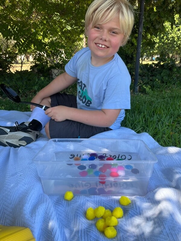 Smiling boy in blue shirt sitting on blanket playing with clear sensory bin containing colorful pompoms outdoors