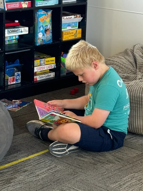 Boy in turquoise shirt sitting cross legged on floor reading colorful picture book near game storage shelves during quiet time