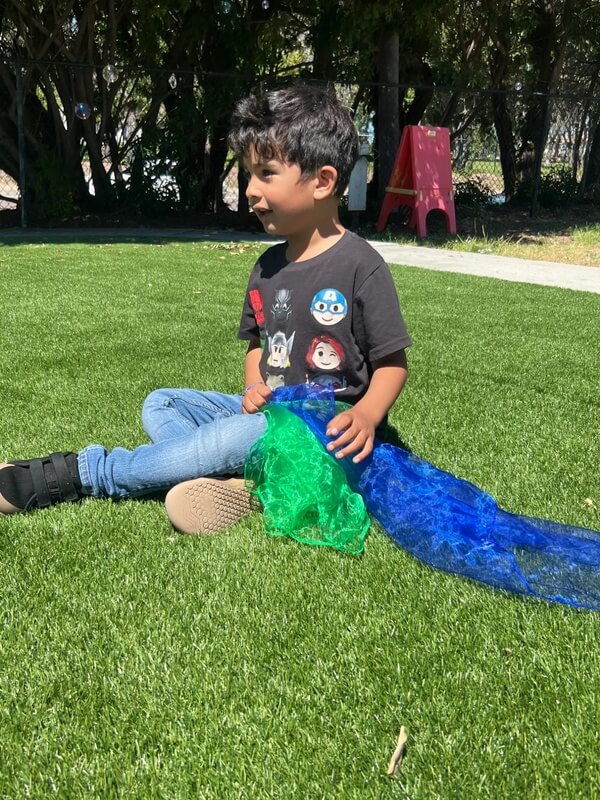 Boy sitting on lawn playing with bright blue and green sensory scarves during outdoor sensory play activity