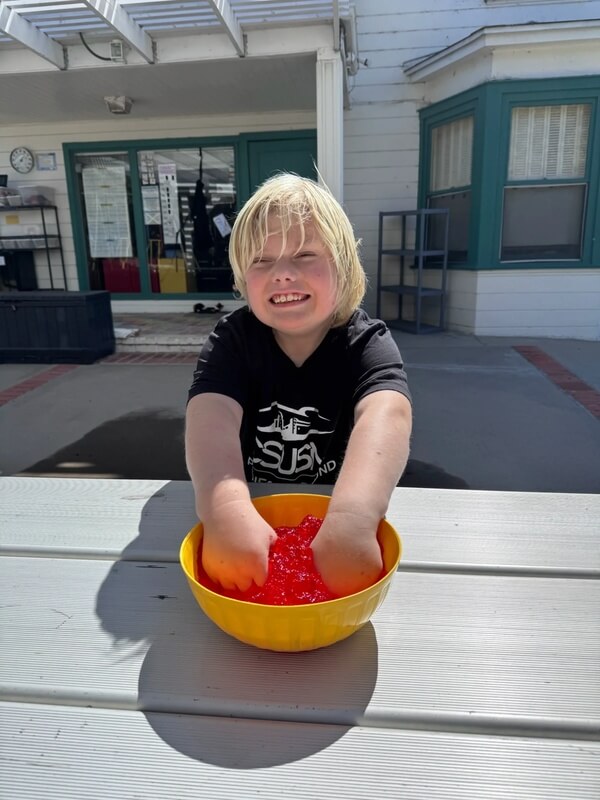 Happy child at outdoor table playing with red translucent sensory slime in yellow bowl during hands-on activity
