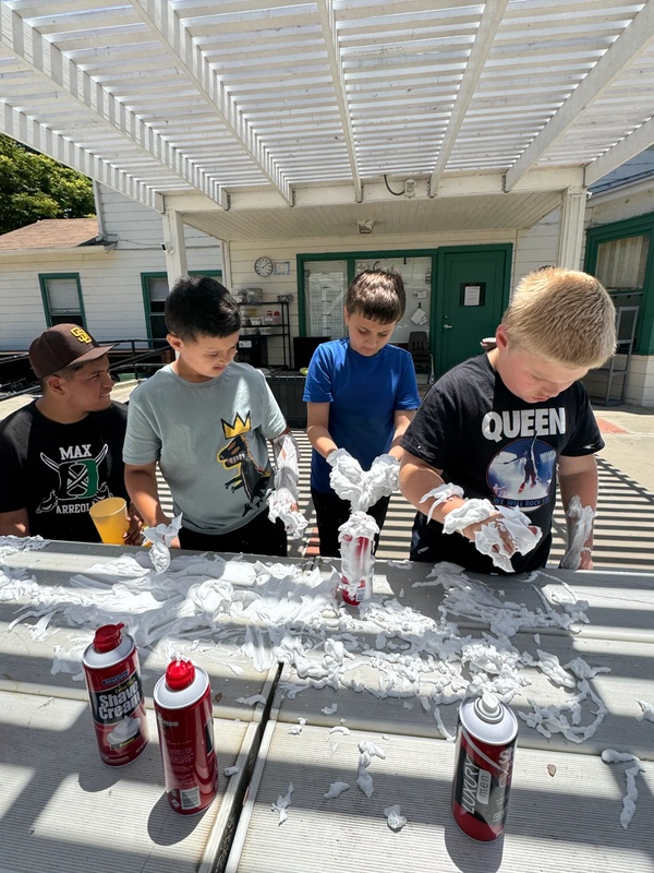 Four children gathered around outdoor table conducting shaving cream eruption experiment with cans and bottles