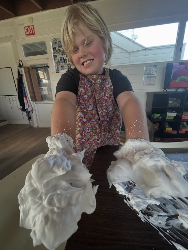 Smiling child with hands covered in white fluffy shaving cream exploring texture during sensory play activity indoors