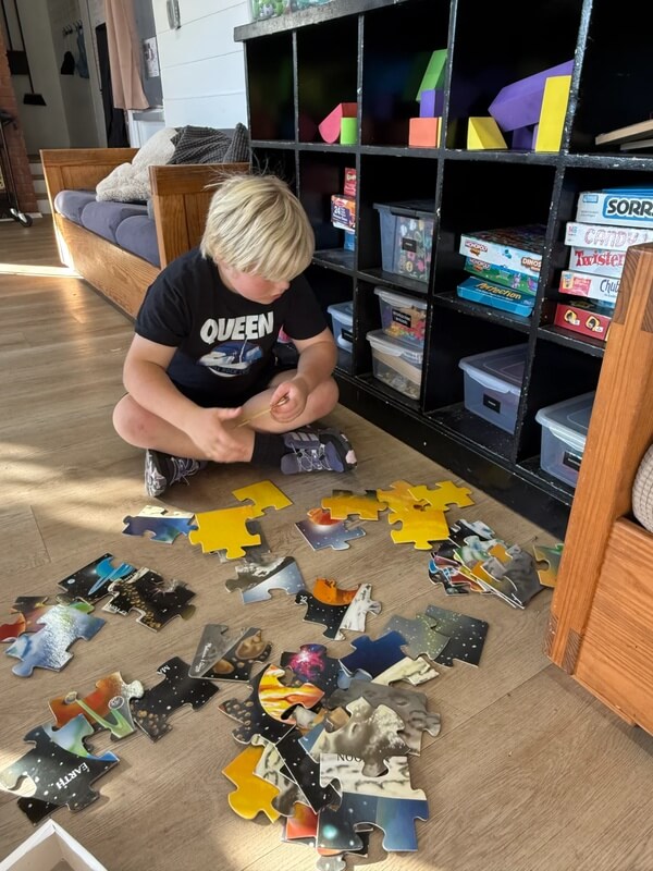 Child in Queen shirt working on large space and planet themed floor puzzle with yellow and dark pieces near organized game storage
