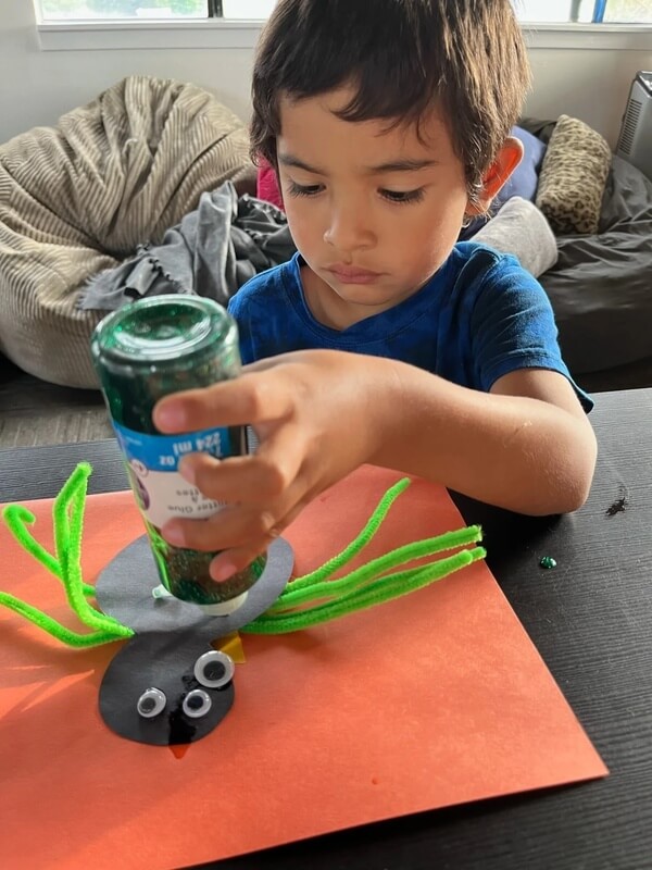 School-age child assembling spider craft using green pipe cleaners, googly eyes, and paint jar at Ivey Ranch program