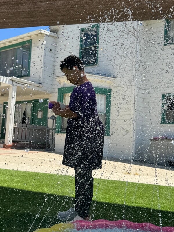 Child standing in outdoor sprinkler spray with water droplets filling the air on sunny day