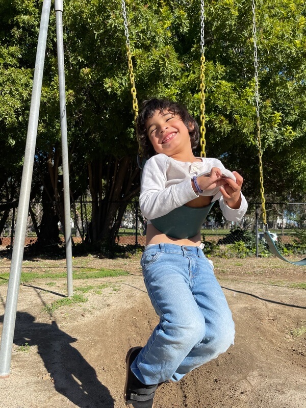 Happy child in white long sleeve shirt swinging on chain swing set with green trees in background