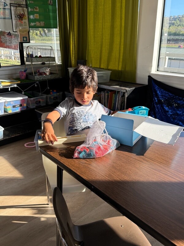 Child working on individual craft project at table with mesh bag and supplies near window during indoor activity time