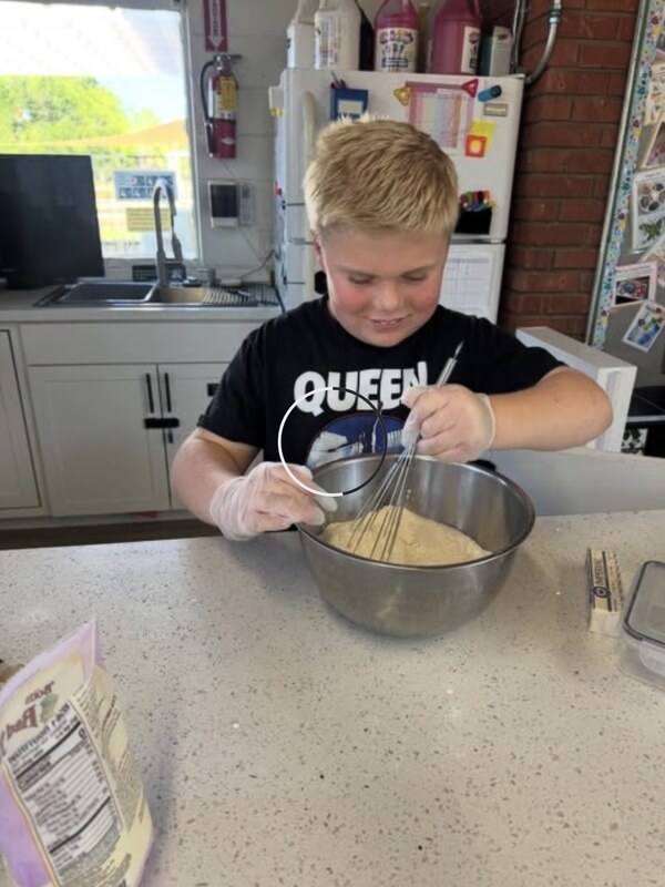 Smiling boy in black Queen shirt using whisk to mix batter in metal bowl in program kitchen with recipe card nearby