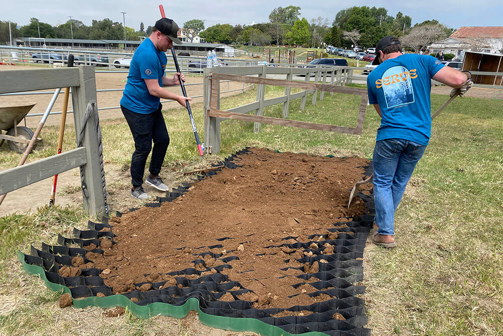 Two volunteers using shovels to prepare and level ground near the arena fence during a service project at Ivey Ranch Park Association in Oceanside, California