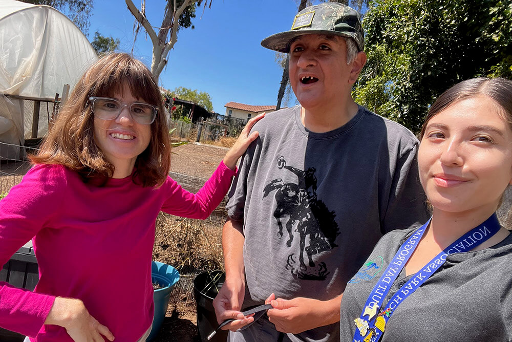 An Ivey Ranch staff member and volunteer smile with an Adult Day Program participant during an outdoor gardening activity on the ranch grounds.