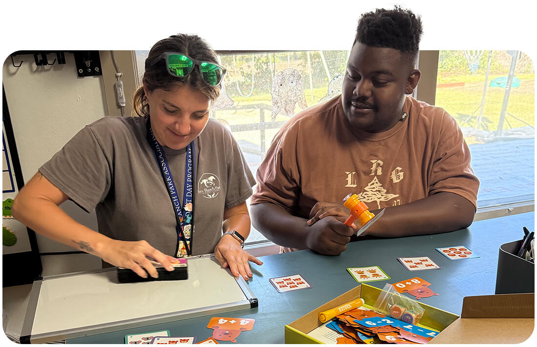 An Ivey Ranch Adult Day Program staff member works one-on-one with a young adult participant on a math card game at an indoor table.