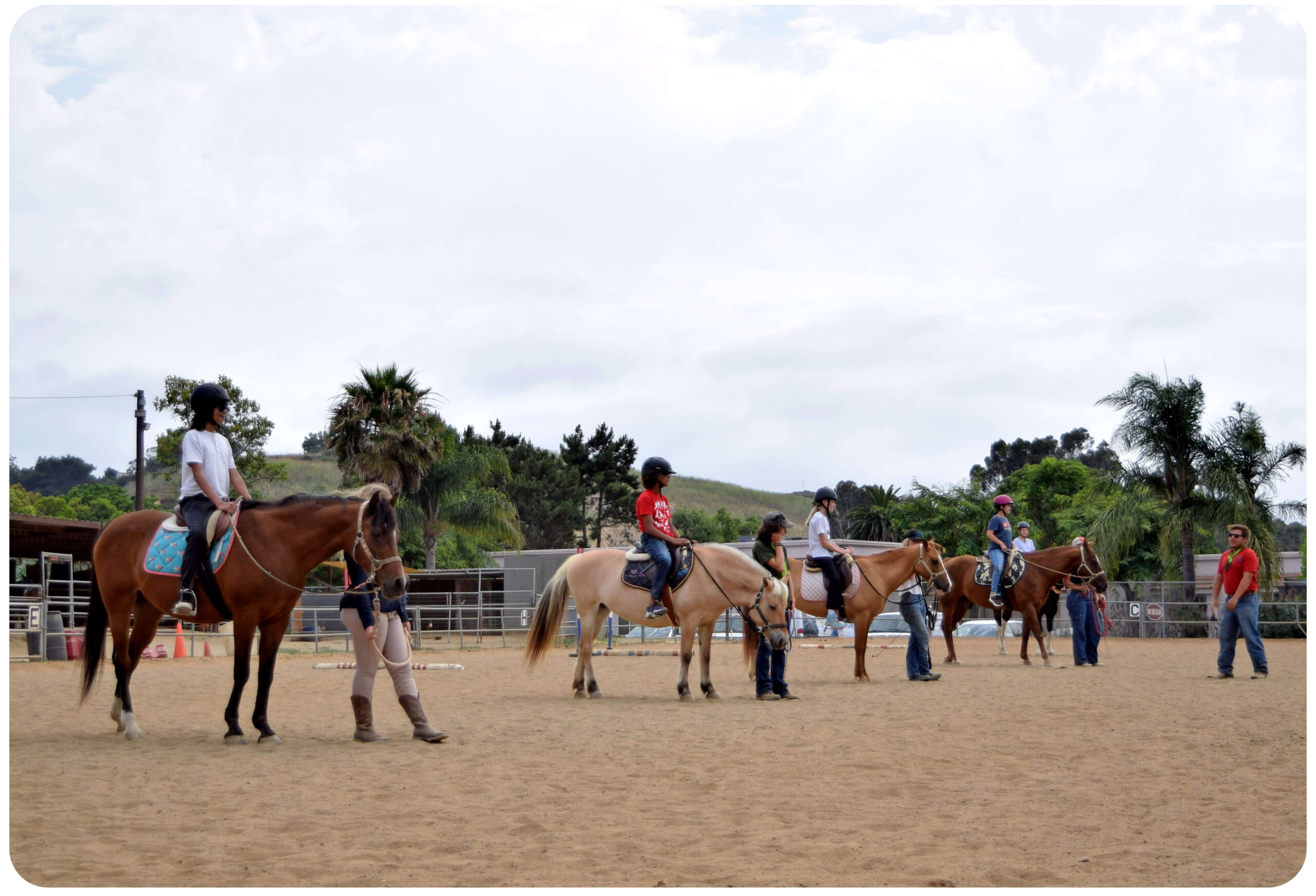 A group of children on horseback line up in the Ivey Ranch arena during a birthday party riding lesson, with instructors guiding them on a cloudy day in Oceanside, CA.