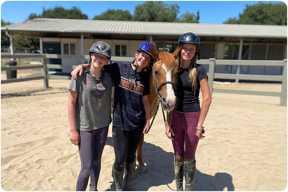 Three smiling kids wearing riding helmets pose together with a chestnut horse in the Ivey Ranch arena during a birthday party experience in Oceanside, CA.