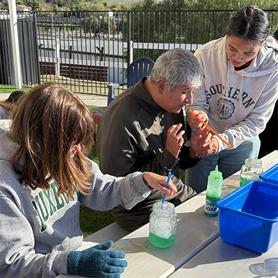 Two Ivey Ranch Care Program volunteers assist an adult participant with a bubble science experiment at an outdoor table.