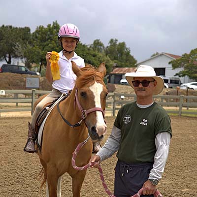 Young rider holding a ribbon on horseback with a volunteer at an Ivey Ranch Park Association Horse Show in Oceanside CA