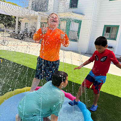 Three children with special needs laugh and play in a splash pad on the Ivey Ranch grounds during a School-Age Program activity day.