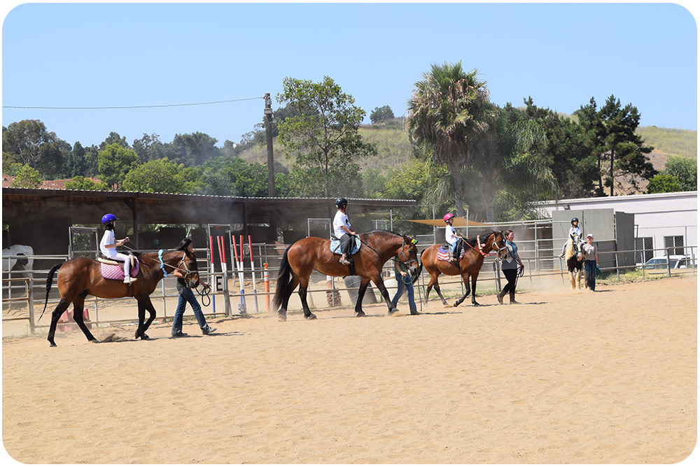A line of young riders on horseback with side walkers make their way around the Ivey Ranch arena during a Scout Program session in Oceanside, CA.