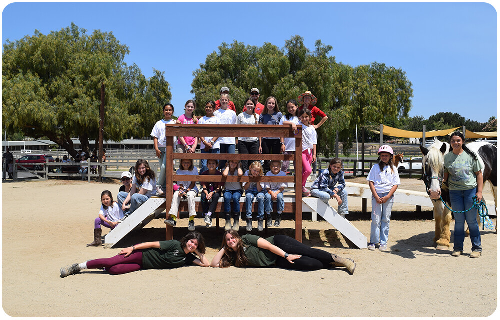 A large group of Summer Horse Camp campers and staff pose together on and around a wooden mounting structure in the Ivey Ranch arena, with a horse standing nearby on a sunny day in Oceanside, CA.