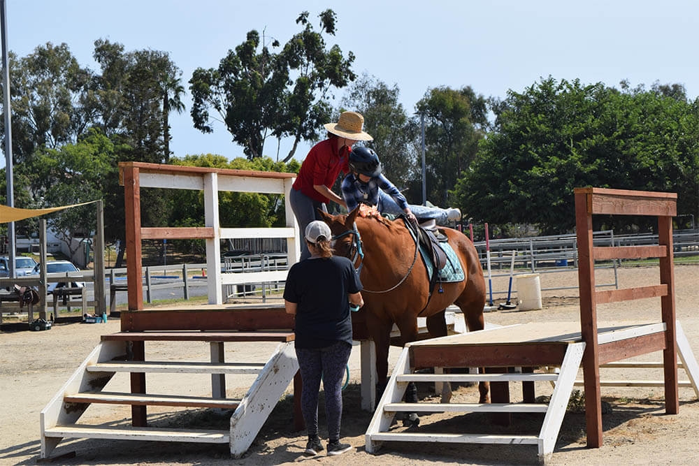 A riding instructor assists a therapeutic riding participant at the mounting ramp at Ivey Ranch Park Association in Oceanside, CA